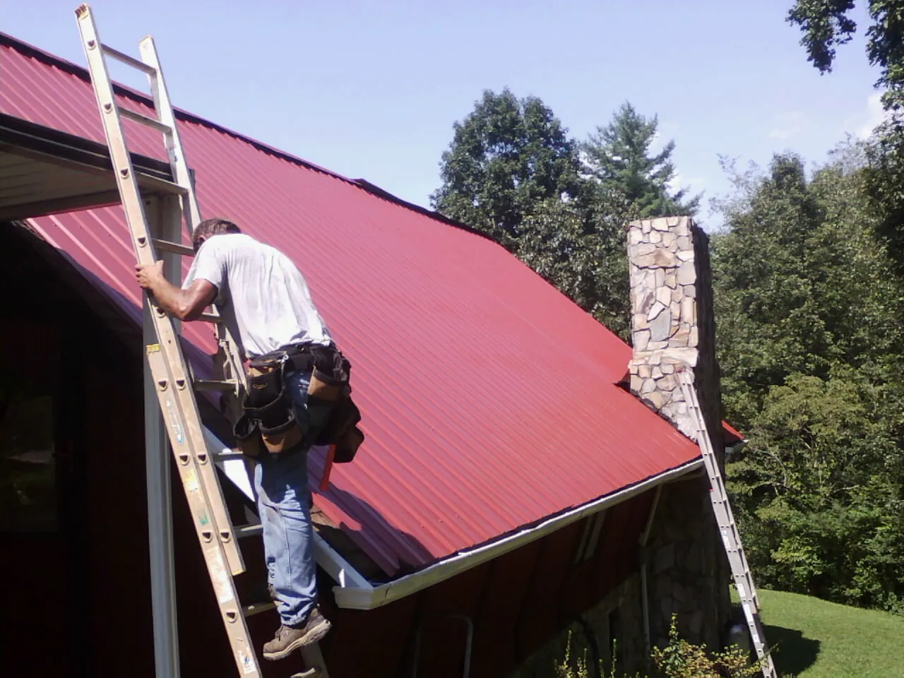 Roofer on a ladder beside a red metal roof and stone chimney — Soffit & Fascia Repair by Roofing Brothers in Parlin